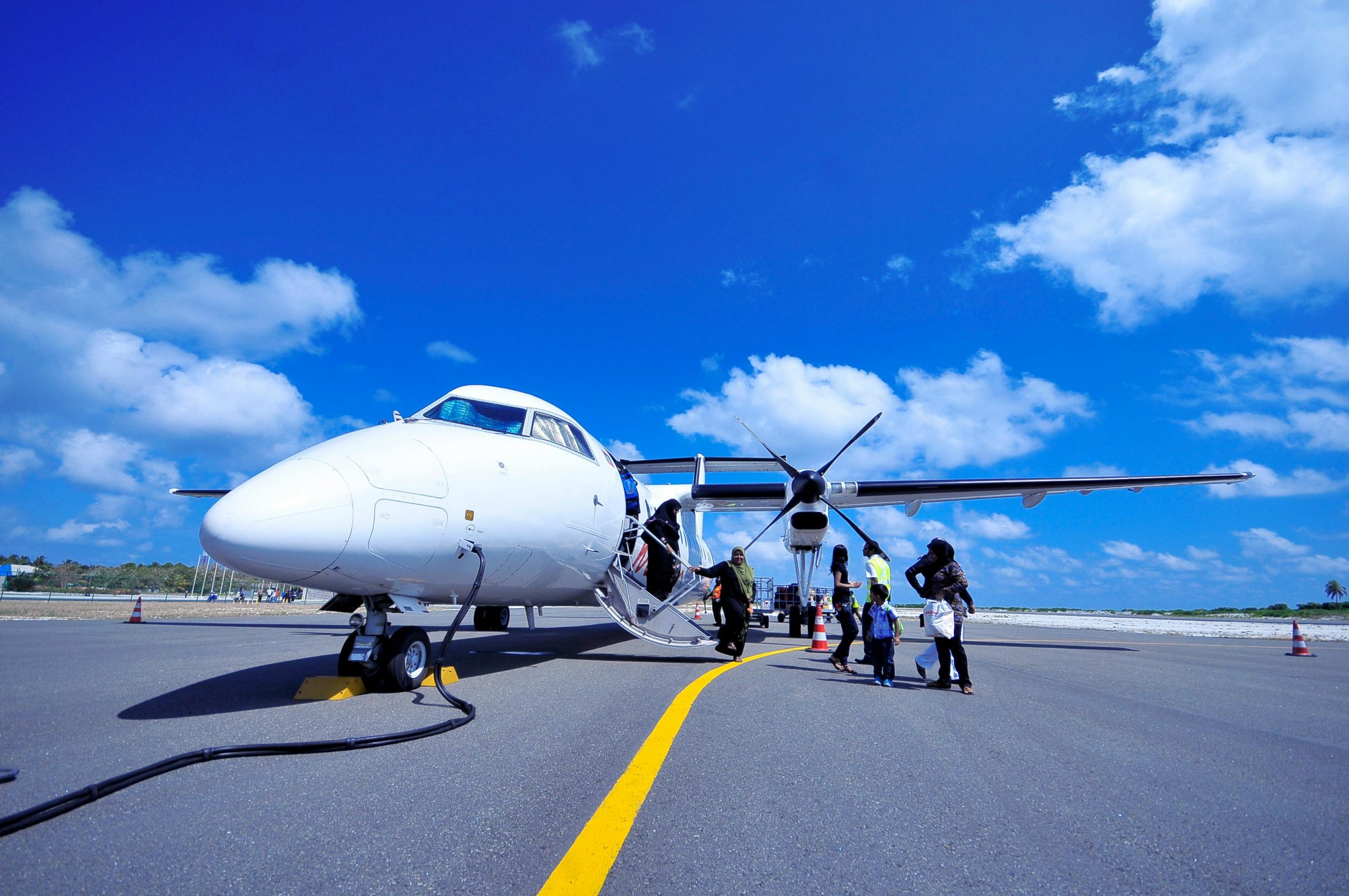 A white private jet with a propeller stands on the tarmac under a bright blue sky with scattered clouds. A group of passengers, including women and children, are seen boarding the aircraft via a small staircase. A ground crew member in a high-visibility vest assists them. The setting appears to be a tropical or remote airport, with palm trees visible in the background.