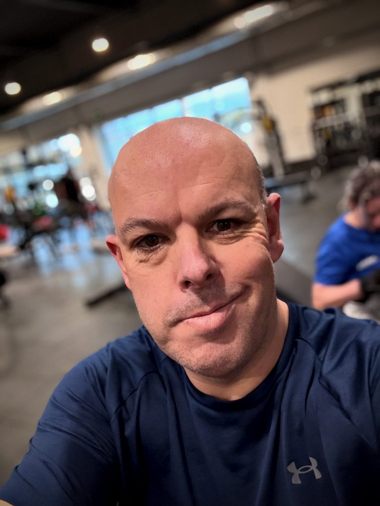 Man taking a post-workout selfie at the gym during his journey of life after Mounjaro, wearing a navy Under Armour shirt with gym equipment in the background