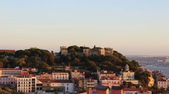 Panoramic view of Lisbon at golden hour, with São Jorge Castle on the hilltop surrounded by trees and colourful houses overlooking the river.