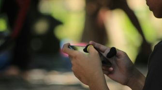 Close-up of someone holding a smartphone and gaming with finger sleeves, with a blurred outdoor background.