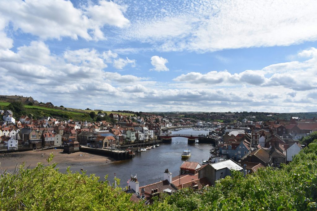 Whitby harbour viewed from above on a bright day, showing the River Esk, colourful rooftops, fishing boats, and the historic bridge under a dramatic cloud-filled Yorkshire sky.