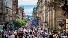 Busy pedestrianised Glasgow city-centre street lined with historic buildings, shops, restaurants and outdoor seating on a sunny day.